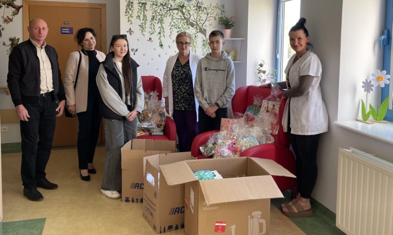 Grupa osób: dorośli i dzieci stoi obok siebie. Osoby znajdują się w pomieszczeniu 