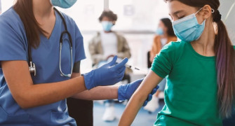 Kobieta i dziewczynka. Obie w maseczkach. Kobieta podaje dziewczynce szczepionkę 