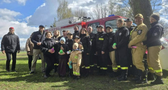 Grupa osób: kobiety, mężczyźni i dzieci stoi obok siebie. Za osobami znajduje się wóz strażacki 