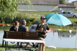 Grupa osób siedzi obok siebie na ławce. Ławka znajduje się przy stawie 