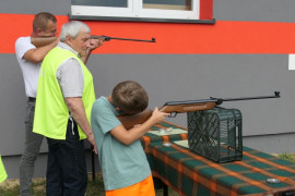 Trzy osoby. Mężczyzna i dziecko strzelają do tarczy z długiej broni. Pomiędzy nimi stoi sędzia 