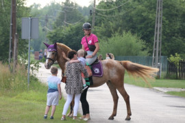Dziewczynka siedzi na koniu. Obok stoi kobieta, która trzyma na ręku małe dziecko. Obok kobiety stoi chłopiec