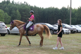 Dziewczynka w różowej bluzce i czapce na głowie siedzi na koniu. Obok stoi dziewczynka w sportowym stroju 