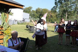 Grupa osób - kobieta w ludowym stroju siedzi na krześle, widoczna jest też osoba w stroju lajkonika oraz dziewczynki w kolorowych strojach 