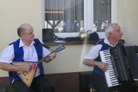 Dwaj panowie w białych koszulach i niebieskich kamizelkach. Siedzą na krzesłach i grają na instrumentach 