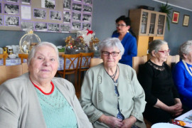 Trzy kobiety siedzą przy stole. Tuż za nimi znajduje się kolejna kobieta