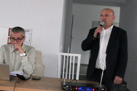 Dwaj mężczyźni. Jeden siedzi przy stole. Drugi stoi i przemawia