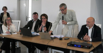 Pięć osób. Cztery osoby siedzą przy stole, jedna przemawia. Na stole stoją laptopy 