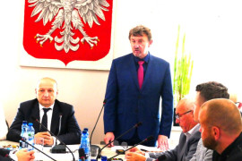 Dwóch mężczyzn w garniturach za stołem prezydialnym. Tuż za nimi na ścianie znajduje się godło Polski 
