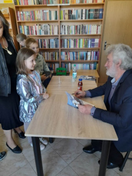 Mężczyzna siedzi przy stoliku. Naprzeciwko niego stoi dziewczynka. Mężczyzna trzyma w ręku książkę i rozmawia z dziewczynką