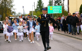 Mężczyzna w mundurze idzie defiladowym krokiem. Za nim idą dziewczynki w białych spódniczkach