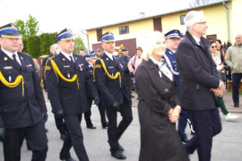 Trzech mężczyzn w strażackich mundurach idzie ulicą w defiladzie. Przed nimi idzie kobieta i mężczyzna 