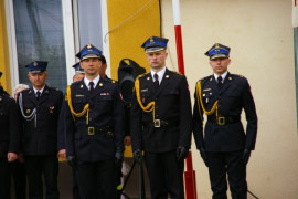 Trzech mężczyzn w mundurach strażackich stoi przy maszcie flagowym