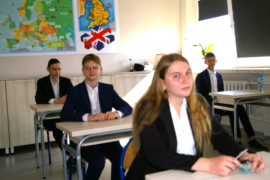 Uczniowie, na pierwszym planie uczennica w galowym stroju siedzą w ławkach w sali egzaminacyjnej  