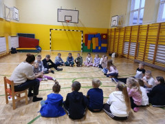 Grupa dzieci siedzi w kręgu w sali gimnastycznej, kobieta prowadzi zajęcia