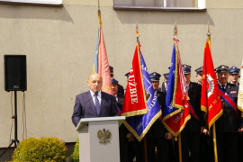 Mężczyzna w garniturze przemawia z mównicy. Za nim stoją poczty sztandarowe