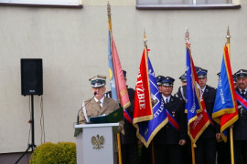 Mężczyzna w mundurze przemawia z mównicy. Za nim stoją poczty sztandarowe