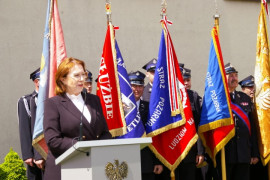 Kobieta przemawia z mównicy. Za nią stoją poczty sztandarowe