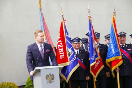 Mężczyzna przemawia z mównicy. W tle poczty sztandarowe