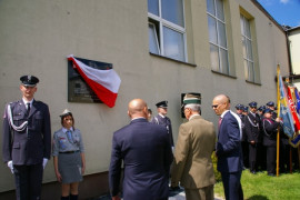 Trzech mężczyzn stoi przed tablicą wiszącą na ścianie budynku i przesłoniętą biało-czerwoną wstęgą. Obok tablicy stoi mężczyzna i kobieta w mundurze