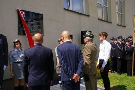 Czterech mężczyzn stoi przed tablicą znajdującą się na ścianie budynku. Z tablicy zwiewa biało-czerwona wstążka