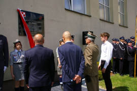 Przed tablicą znajdującą się na ścianie budynku stoi czterech mężczyzn, jeden z nich jest w mundurze