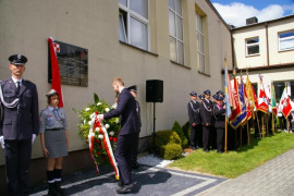 Mężczyzna składa biało - czerwony wieniec kwiatów pod tablicą znajdującą się na ścianie budynku. Obok tablicy stoją mężczyzna i kobieta w mundurach