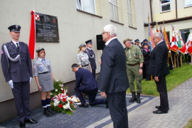 Dwóch mężczyzn stoi przed tablicą. Jeden mężczyzna składa kwiaty. Obok tablicy stoją osoby w mundurach