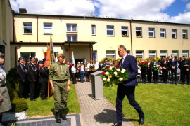 Mężczyzna w garniturze składa wiązankę kwiatów pod tablicą pamiątkową. W tel widać grupę ludzi 