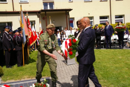 Dwóch mężczyzn i kobieta.  Mężczyzna w garniturze podaje wiązankę biało-czerwonych kwiatów mężczyźnie w mundurze