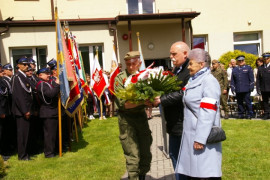 Kobieta i mężczyzna w cywilnych strojach przekazują wiązankę kwiatów mężczyźnie w mundurze. Kobieta na rękawie płaszcza ma biało-czerwoną opaskę