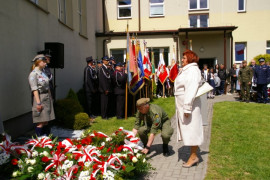 Kobieta w białym płaszczu stoi przed tablicą wiszącą na ścianie budynku. Pod tablicą leżą biało-czerwone wiązanki kwiatów