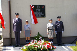 Dwaj mężczyźni w mundurach oraz dwie kobiety w mundurach stoją przy tablicy znajdującej się na ścianie budynku. Pod tablicą leżą wiązanki biało-czerwonych kwiatów 