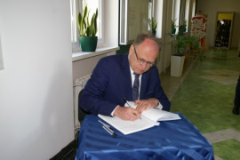Mężczyzna w garniturze i okularach siedząc na krześle wpisuje się do pamiątkowej księgi