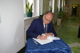 Mężczyzna w garniturze i okularach siedząc na krześle wpisuje się do pamiątkowej księgi