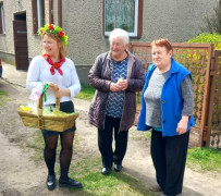 Trzy kobiety stojące przed domem. Jedna z nich trzyma w ręku ozdobiony kwiatami kosz 