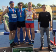 Trzej mężczyźni w strojach sportowych stoją a podium. Obok nich stoi kolejny mężczyzna 