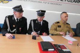 Trzej mężczyźni w mundurach siedzą za stołem. Trwa podpisywanie umowy