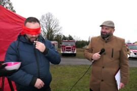 Dwóch mężczyzn - jeden ma zawiązane oczy czerwoną chustką. Obok niego stoi mężczyzna trzymający w ręku mikrofon 