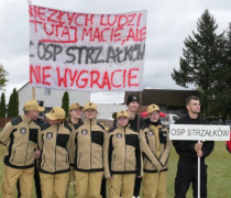 Strażacy stoją obok siebie. Widoczny jest transparent 