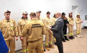 Grupa osób, większość w strojach strażackich. Moment wręczania legitymacji członkowskich. 