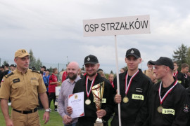 Grupa mężczyzn stojąca obok siebie na murawie boiska. Jeden z nich trzyma w ręku puchar i dyplom 