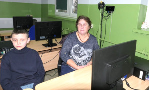 Kobieta i chłopiec siedzą w szkolnej ławce. Przed nimi znajduje się monitor komputerowy 