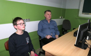 Mężczyzna i chłopiec siedzą w szkolnej ławce. Przed nimi znajduje się monitor komputera 