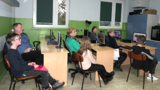Dorośli i dzieci siedzą w szkolnych ławkach, na których stoją monitory komputerów 
