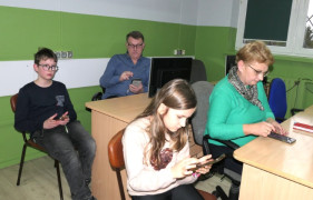 Kobieta, mężczyzna i dwoje dzieci siedzą w szkolnych ławkach. Wszyscy korzystają ze smartfonów 