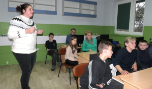 Dorośli i dzieci siedzą w szkolnych ławkach. Po sali chodzi kobieta 