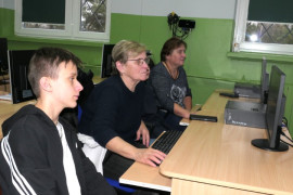 Dwie kobiety i chłopiec siedzą w szkolnej ławce. Na ławce stoi monitor komputera 