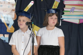 Dziewczynka i chłopiec stoją obok siebie. Oboje mają na głowie granatowe birety 