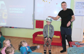 Grupa dzieci i mężczyzna. Mężczyzna prezentuje sprzęt wykorzystywany we wspinaczce górskiej 